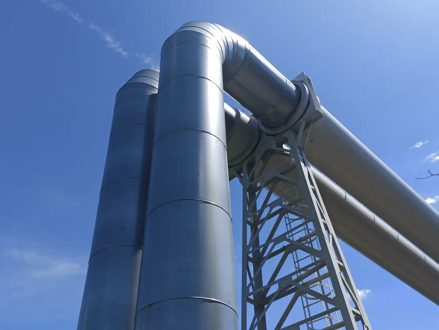 Looking up at large steel industrial pipes and a support gantry against a clear blue sky.